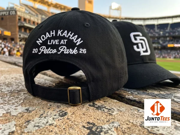 A close-up shot of the back of a black baseball cap featuring white arched embroidery that reads 'NOAH KAHAN LIVE AT 20 Petco Park 26' with an adjustable black strap and brass buckle, set against a blurred stadium background.