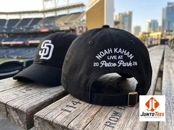 A close-up shot of the back of a black baseball cap featuring white arched embroidery that reads 'NOAH KAHAN LIVE AT 20 Petco Park 26' with an adjustable black strap and brass buckle, set against a blurred stadium background. (Detail view #3)