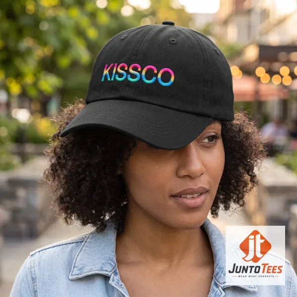 Front view of a black Harry Styles Kissco merchandise cap with rainbow embroidered text on a wooden table. (Detail view #4)